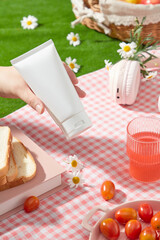 Cosmetics advertising photo with a picnic concept from a high angle, an empty label tube is held by a beautiful hand, below is a pink plaid rug containing some picnic items