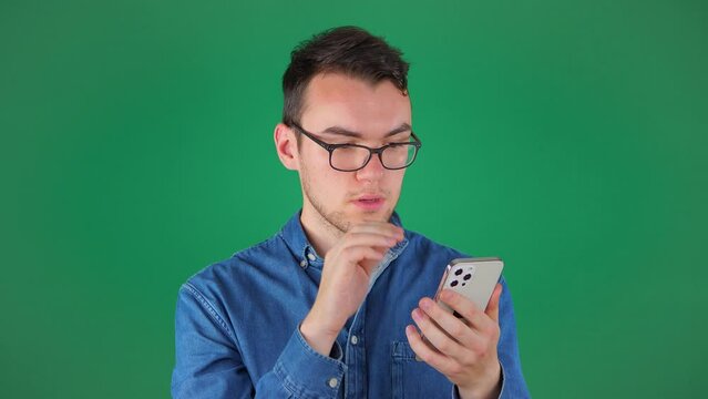 A young man with glasses in a blue shirt talks on the phone, writes SMS messages, corresponds with friends, on a green background. Slow Motion