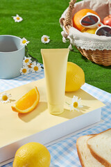 Outside picnic activity landscape, green grass containing a blue carpet with a yellow book, sandwich, citrus fruits and an empty label cosmetic tube in yellow color. High angle view, space for text