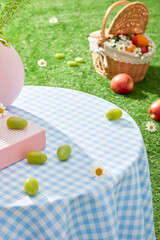 A table with a blue tablecloth, has a few grapes scattered on top. In the distance is a rattan basket containing fruit placed on the grass. Photo of high angle with empty space for displaying