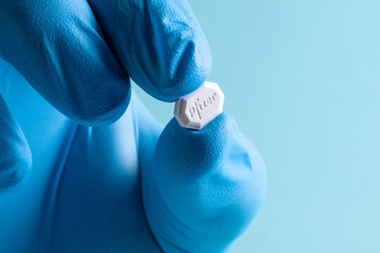 A white PFIZER pill held by a blue-gloved hand against a soft blue background.