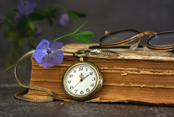 Still life with a pocket watch on a chain, an old, tattered book, a periwinkle flower and glasses.