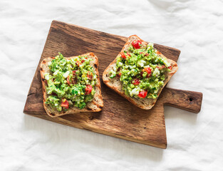 Avocado, egg, red onion, cherry tomatoes, greens spread whole grain grilled bread sandwich on a wooden cutting board on a light background, top view