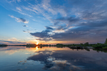 Naklejka premium A breathtaking view of a spring sunset over a calm river, showcasing a picturesque landscape with a mirror-like reflection of the sky.