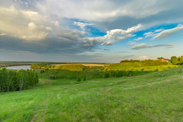 A stunning view of a river valley from a grassy hillside, with lush greenery, fluffy clouds, and a tranquil blue sky. The scene captures the beauty of a spring evening.