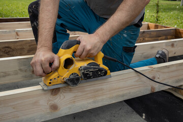 A carpenters hands are shown using a power planer to smooth out a wooden beam. The carpenter is wearing blue pants and is kneeling on the wooden beams.