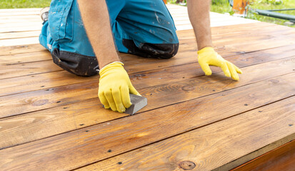 A close-up view of a person wearing yellow gloves sanding a wooden deck with a block sander. The deck is made of weathered wood planks, and the person is kneeling on the deck.
