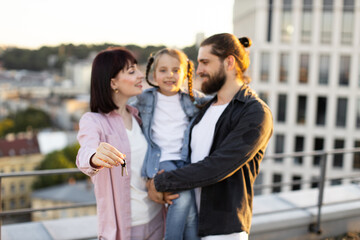 Happy family standing on rooftop, holding keys and celebrating new home with young daughter, representing excitement and togetherness.