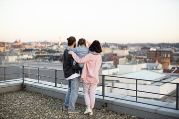 Family of three, embracing and enjoying scenic city view during sunset from rooftop, expressing togetherness and love.