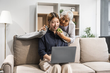 A mature mom and her young Asian daughter sit on a sofa, working with a laptop. The mom supports, encourages, and stays by her side, helping with warmth and caring love on Mother's Day.