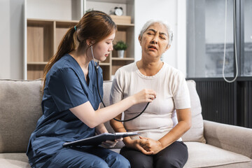 A young female nurse of Asian descent provides care to an elderly patient, a woman, sitting on a sofa.