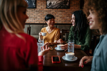 Smiling diverse female friends sit in a cafe laughing and talking over a coffee