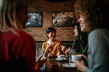 Four Young Female Friends Meeting Sit At a Table In a Coffee Shop And Talk. Focus On A Asian Woman Talking