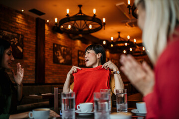 Smiling Asian woman showing her friends what clothes she bought in shopping, they are in the restaurant