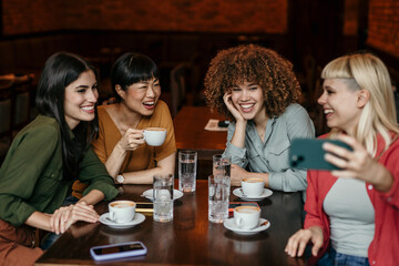 A smiling group of female friends sitting in the cafe, having a casual time and taking a selfie with a phone