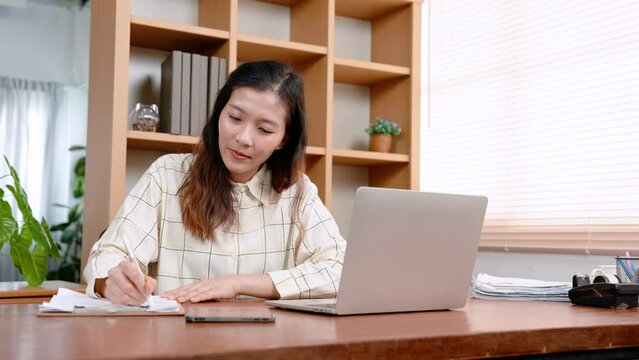 Beautiful Asian office worker, sitting working on laptop computer placed on table in front, with carefree expression, reached out hand picked up pen write down main points work in notebook.