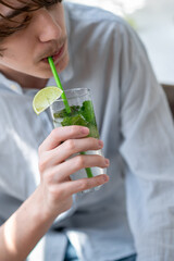 Teenage boy in light blue shirt drinking refreshing summer iced cocktail, holding glass with mojito...