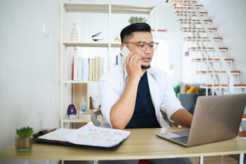 Asian male freelancer making a phone call, share good news about project working at home office. Remote job concept.