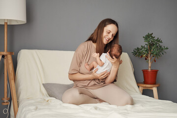 Beautiful Caucasian mother dressed in beige attire sitting on the sofa at home, cradling her...