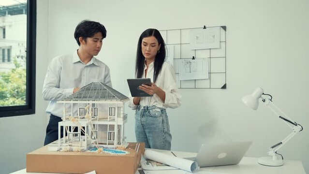 Beautiful young caucasian project manager brief asian engineer by using tablet while engineer measuring house model on meeting table with blueprint, architectural equipment scatter around. Immaculate