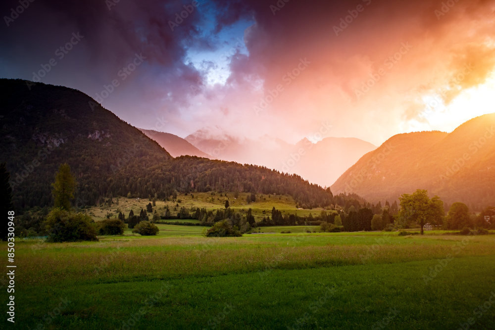 Fototapete Majestic view of green fields with hills under a colorful cloudy sky. Julian Alps, Slovenia, Europe.