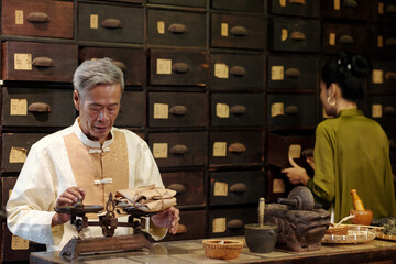 Senior pharmacist measuring package with herbs with his assistant working in background