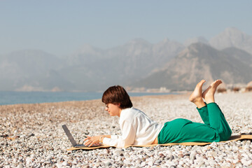 Digital nomad woman traveling the world working with laptop, the girl works on the beach, spends a working day on the beach against the background of mountains and the sea