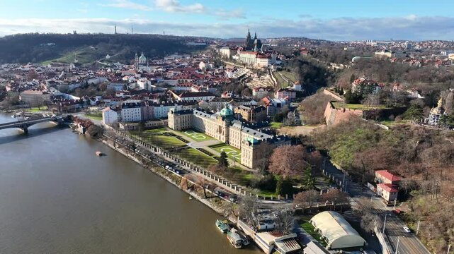 Prague, Czechia, December 2022. Drone gradually orbits the The Straka Academy on the banks of the Vltava river with Prague Castle and Petrin hill in the distance.
