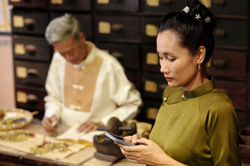 Asian assistant texting message on her smartphone while apothecary working in background in drug store
