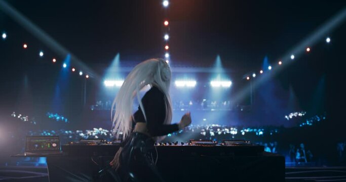 Female DJ Delivers a Live Performance of Electronic Music while Standing Behind Turntable Decks at a Massive Concert. Colorful Lights and Dance Beats Play at an EDM Concert. Footage from the Back