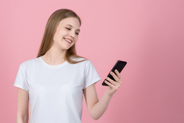 Attractive caucasian young blonde woman in casual white t-shirt smiling and using mobile phone isolated on pink studio background. Wireless, app, internet, social media, online.