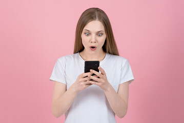 Surprised attractive caucasian young blonde woman in casual white t-shirt using mobile phone isolated on pink studio background. Wireless, social media, internet, app.