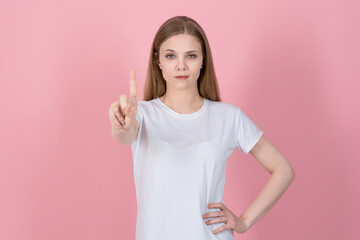 Attractive serious caucasian young blonde woman in casual white t-shirt showing stop, one finger gesture, taboo sign isolated on pink studio background.