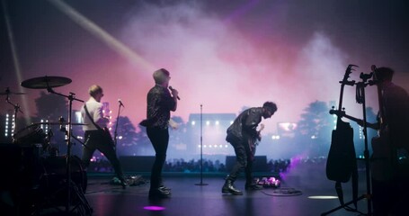 Rock Music Concert at a Festival: Energetic Indie Band Performs Live on Open Air Stage at Sunset, Using Instruments to Play Indie, Punk, and Alternative Tunes. Cinematic Footage From the Back