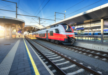 Fototapeta premium High-speed red passenger train at railway station platform under clear blue sky at sunset. Train station. Modern railway transportation concept. Railroad. Commercial. Urban rail transport in Austria