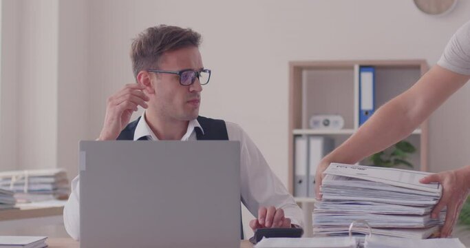 Frustrated businessman calculating, watching laptop screen, feeling annoyed with extra paper work pile. Office overworked manager in fatigue, exhaustion, great stress, overburdened worn out accountant