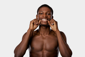A shirtless African American man diligently flossing his teeth in a white studio setting.