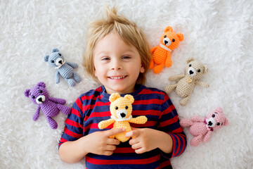 Little toddler child, cute blond boy, lying down in bed with many teddy bears, handmade amigurumi...