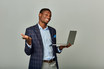 Young African American businessman in checkered blazer confidently holds a laptop on grey background.