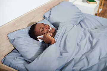 A young African American man laying in bed, holding a smartphone in his hand as he wakes up in the morning.