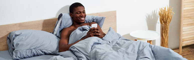 A young African American man lying in bed, fully engaged with his cell phone in the morning.