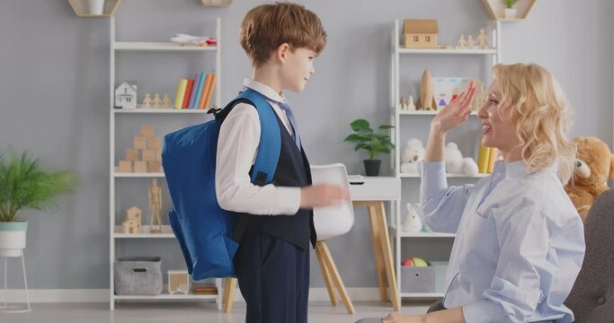 Mother shows love, care and support to child on first day of primary school. Mom gives high five, hugs and says goodbye to son in uniform with backpack as he leaves nursery room before lessons start