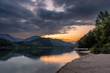 A picturesque scene at Almsee Lake in Austria, featuring a calm lake reflecting a golden sunset, with a small cabin nestled in the water and towering mountains in the background. 