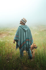 Young man with hat and poncho, posing with his dog in a foggy meadow.