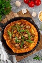 Savory Dutch baby pancake with cheese, fried mushrooms and arugula in cast iron pan on a concrete background. Vegetarian breakfast.