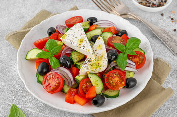 Greek salad of fresh juicy vegetables, feta cheese, herbs and olives on a white plate on a light concrete background. Healthy food. Copy space, top view.