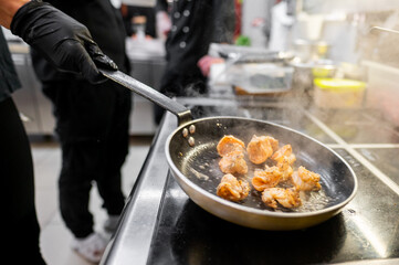 Close-up of a frying pan on a stove, sautéing chunks of shrimp with steam rising. A person in black gloves holds the handle.