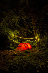A red tent illuminated by fairy lights nestled amongst trees in a dark forest.