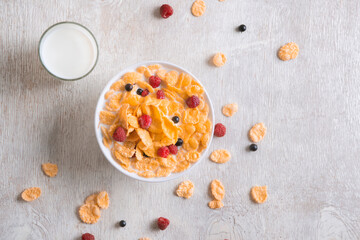 bowl of corn granola with milk, fresh raspberries, blueberries . milk in a jug and a glass  on white wooden board for healthy breakfast, top view