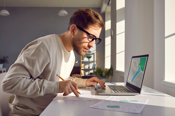 Portrait of professional cartographer looking at laptop monitor screen, working with printed...
